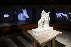 a marble sculpture of a horse in the fron, behind video works of horses in the back wall